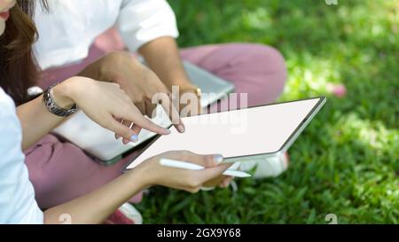 Zugeschnittenes Bild von zwei Freundinnen sitzt in einem Stadtpark und schaut zusammen auf einem digitalen Tablet-Bildschirm. Arbeits- und Entspannungskonzept Stockfoto