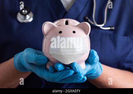 Doktor oder Krankenschwester tragen chirurgische Handschuhe Holding Piggy Bank trägt medizinische Gesichtsmaske. Stockfoto