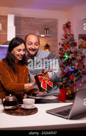 Glückliches Paar zeigt Geschenk an entfernte Freunde während Online-Videocall-Meeting auf Laptop-Computer. Fröhliche lächelnde Familie genießen Wintersaison feiert weihnachtsfeiertage in Weihnachten dekoriert Küche Stockfoto