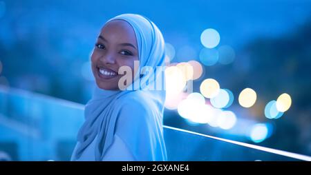 Afrikanisch-muslimische Frau in der Nacht auf dem Balkon lächelt vor der Kamera mit Bokeh-Lichtern im Hintergrund Stockfoto