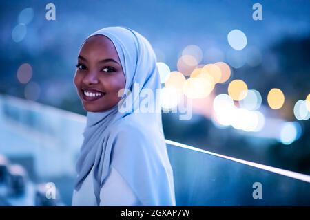 Afrikanische Moderne muslimische Frau in der Nacht am Balkon lächelnd an Kamera mit Stadt bokeh Lichter im Hintergrund Stockfoto
