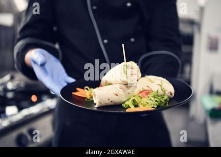 Koch tragen Gesicht schützende medizinische Maske für den Schutz vor Corona-Virus-Krankheit mit Teller mit authentischen Sandwich im professionellen Restaurant KI Stockfoto