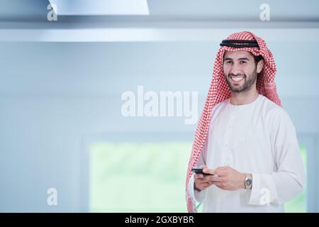 Junge moderne muslimische arabische Geschäftsmann in traditioneller Kleidung, während Smartphone zu Hause Stockfoto