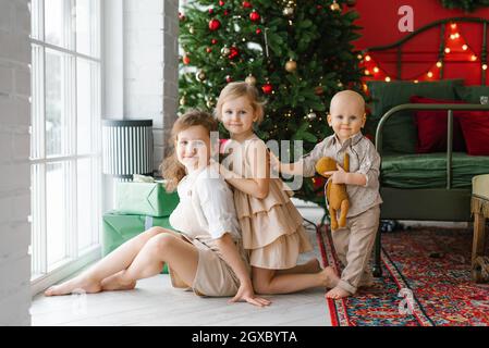 Drei Kinder, ein Junge und Mädchen sitzen vor dem Hintergrund eines Weihnachtsbaums im Schlafzimmer am Fenster und bereiten sich auf das neue Jahr vor Stockfoto