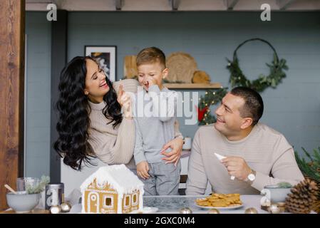 Eine glückliche europäische oder amerikanische Familie mit einem kleinen Kind steht zu Weihnachten am Küchentisch und schmückt hausgemachte Kuchen mit Weihnachtsgingerbrea Stockfoto