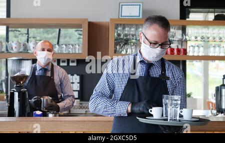 Kellner in einer medizinischen Schutzmaske serviert den Kaffee im Restaurant Durin Coronavirus Pandemie, die neue normale Konzept Stockfoto