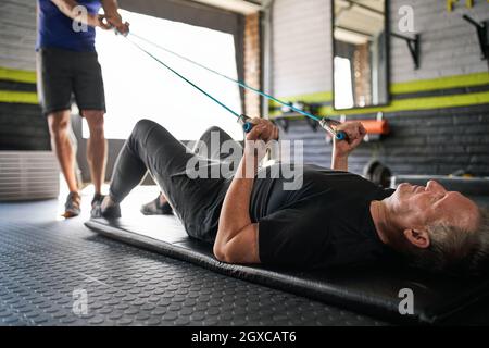 Männlicher Personal Trainer hält sich an elastischen Gummiwiderstandbändern, während ein älterer Mann auf mattem Boden liegt und Bizeps für Stärke A macht Stockfoto