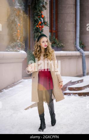 Eine junge Hipsterfrau, die durch die schneebedeckten Straßen der Stadt geht, hält einen eingetopften Weihnachtsbaum in ihren Händen. In einem burgunderroten Rock und einem beigen Mantel Stockfoto