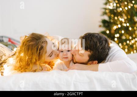 Glückliche Mama und Papa liegen auf dem Bett neben ihrem Kind vor dem Hintergrund der Weihnachtsbeleuchtung und küssen ihn auf die Wangen Stockfoto