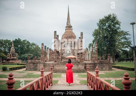 Die Szenerie einer Frau mit ihrem Regenschirm im Lanna-Stil, der an einem bewölkten Tag in der Regenzeit in der Sukhothai Provinc vor dem Tempel Wat Mahathat steht Stockfoto