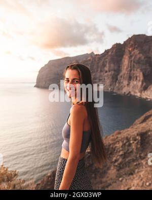 Seitenansicht eines lächelnden Weibchens mit langen Haaren auf einem Kamm, das beim Sonnenuntergang auf Teneriffa in Spanien die Kamera gegen den Ozean ansah Stockfoto