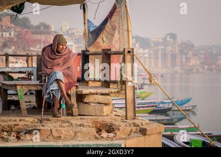 INDIEN, VARANASI - 27. NOVEMBER 2015: Ethnischer Mann sitzt auf einer Holzbank in der Nähe eines Bootes am Flussufer in Indien Stockfoto