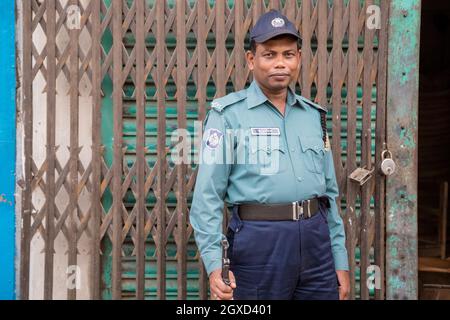 INDIEN, BANGLADESCH - 6. DEZEMBER 2015: Ethnisch bewaffneter Mann in Polizeiuniform und Mütze, der in der Nähe von Metalltoren eines verwitterten Gebäudes steht und schaut Stockfoto