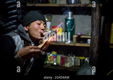 NEPAL, HIMALAYA - 6. NOVEMBER 2015 Junge ethnische Männer in legerer Kleidung und Hut rauchen Zigarette, während sie am Tisch im Café sitzen und die Kamera betrachten Stockfoto