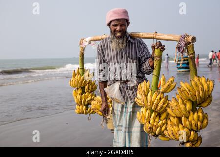 INDIEN, BANGLADESCH - 4. DEZEMBER 2015: Ethnischer Bauer in legerer Kleidung, der am Strand mit frischen Bananen unterwegs ist und dabei die Kamera anschaut Stockfoto