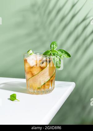Ein Glas kalten Cocktails mit Gurkenscheibe und Basilikumblättern auf einem weißen Tisch mit grünem Hintergrund Stockfoto