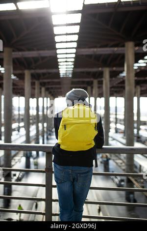 Rückansicht eines nicht erkennbaren männlichen Reisenden mit Rucksack, der in der Nähe des Geländers auf der Passage über Zügen am Bahnhof stand Stockfoto