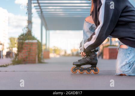 Seitenansicht einer kurzgeschnittenen, nicht erkennbaren jungen Frau, die einen schwarzen Pullover und eine hellblaue Jeans trägt, mit Schlitzen, die Rollerblätter auf einem Knie aufknöpfen Stockfoto