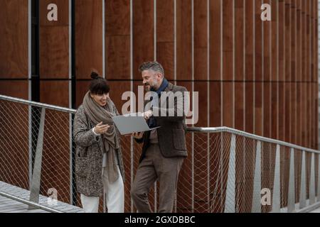 Ein gutaussehender Geschäftsmann mit einer Kollegin, die einen Laptop benutzt und auf einer Terrasse vor dem Büro lächelt und arbeitet. Grauhaariger Mann im Herbst braun trendigen Stil Stockfoto