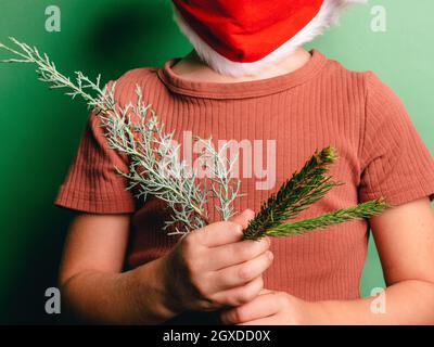 Kurzer unerkennbarer Junge in rotem Weihnachtsmann Hut und Maske zum Schutz vor COVID mit Tannenzweigen Stockfoto