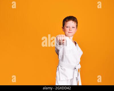 Selbstsicherer Junge in weißem Kimono, der Karate Punch macht und mit lächelnd vor orangefarbenem Hintergrund auf die Kamera blickt Stockfoto
