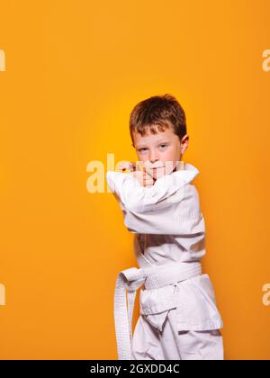 Seriöser Junge mit weißem Kimono, der vor einem leuchtend orangefarbenen Hintergrund karate Haltung einnimmt und die Kamera anschaut Stockfoto