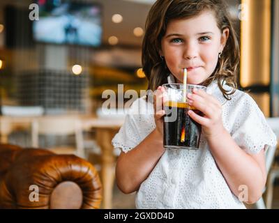 Crop charmante Mädchen trinken gesüßte kohlensäurehaltige Erfrischungsgetränk aus Glas mit Stroh und Blick auf die Kamera Stockfoto