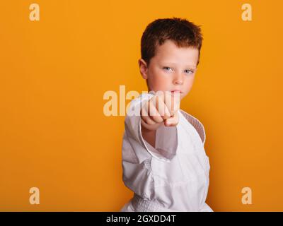 Selbstsicherer Junge in weißem Kimono, der Karate Punch macht und mit ernstem Blick vor orangefarbenem Hintergrund auf die Kamera blickt Stockfoto