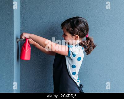 Seitenansicht des kleinen Mädchens, das in der Nähe der grauen Wand steht und tagsüber Wasser in die Gießkanne gießt Stockfoto