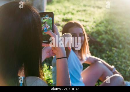 Schneiden Sie nicht erkennbare weibliche Teenager Foto von lächelnden besten Freundin auf dem Handy, während Sie Zeit auf der Wiese in Hinterleuchtung Stockfoto