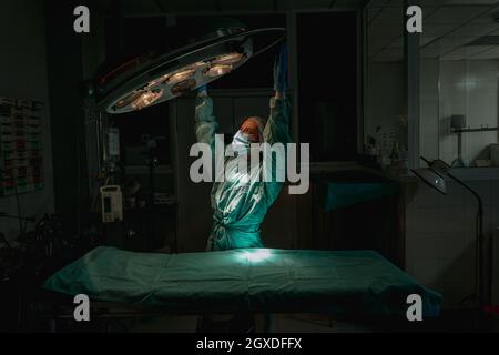 Junge weibliche Tierärztin in einer chirurgischen Uniform und sterilen Maske, die nach oben schaut, während sie die Lampe über dem medizinischen Textil in der Klinik justiert Stockfoto