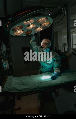 Junge weibliche Tierärztin in einer chirurgischen Uniform und sterilen Maske, die nach unten schaut, während sie die Lampe über dem medizinischen Textil in der Klinik justiert Stockfoto