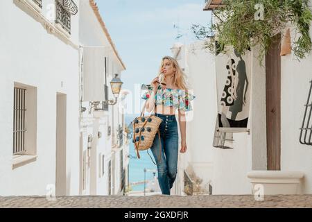 Verträumte Hündin mit Weidensack und Sonnenbrille in der Hand, die auf einer gepflasterten Straße zwischen weißen Häusern in der Küstenstadt unterwegs ist Stockfoto