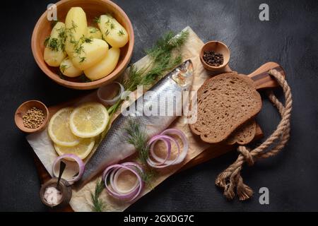 Heringsfisch auf Holzbrett mit Pfeffer, Kräutern, roter Zwiebel und Zitrone auf schwarzem Hintergrund. Draufsicht mit Kopierraum mit Kartoffelscheiben und Roggenfarbe Stockfoto