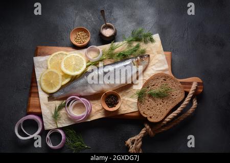 Heringsfisch auf Holzbrett mit Pfeffer, Kräutern, roter Zwiebel und Zitrone auf schwarzem Hintergrund. Draufsicht mit Kopierraum mit Kartoffelscheiben und Roggenfarbe Stockfoto