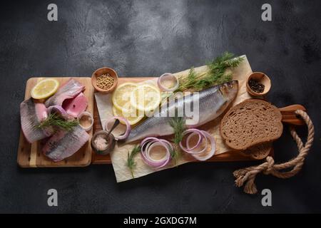 Heringsfisch auf Holzbrett mit Pfeffer, Kräutern, roter Zwiebel und Zitrone auf schwarzem Hintergrund. Draufsicht mit Kopierraum mit Kartoffelscheiben und Roggenfarbe Stockfoto