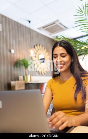Lächelnde junge Unternehmerin, die am Tisch sitzt und Netbook durchstöbert, während sie am modernen Arbeitsplatz arbeitet Stockfoto