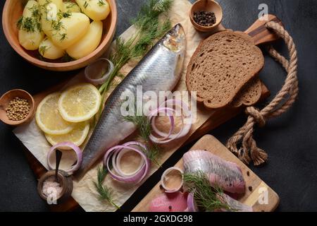 Heringsfisch auf Holzbrett mit Pfeffer, Kräutern, roter Zwiebel und Zitrone auf schwarzem Hintergrund. Draufsicht mit Kopierraum mit Kartoffelscheiben und Roggenfarbe Stockfoto