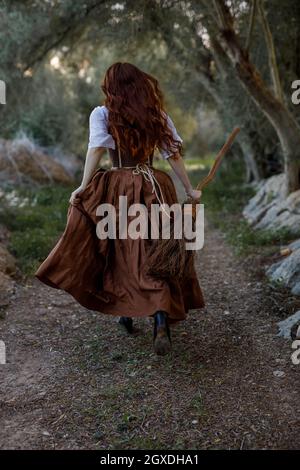 Rückansicht einer unerkennbaren Hexe in Kleid und mit Broomstick, der im Herbstwald entlang des Weges läuft Stockfoto