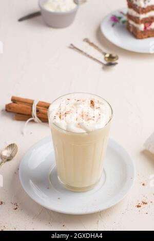 Ein Glas Milchpunsch mit Zimtpulver auf geschlagenem Eiweiß gegen ein Kuchenstück auf dem Cafeteria-Tisch auf hellem Hintergrund Stockfoto