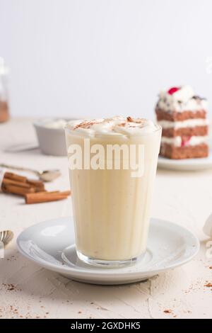 Ein Glas Milchpunsch mit Zimtpulver auf geschlagenem Eiweiß gegen ein Kuchenstück auf dem Cafeteria-Tisch auf hellem Hintergrund Stockfoto