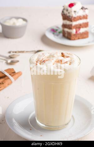 Ein Glas Milchpunsch mit Zimtpulver auf geschlagenem Eiweiß gegen ein Kuchenstück auf dem Cafeteria-Tisch auf hellem Hintergrund Stockfoto
