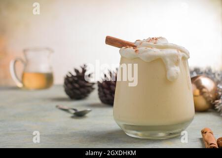 Glas Milchpunsch mit Zimtpulver auf geschlagenen Eiweiß gegen Pinienzapfen am Weihnachtstag auf hellem Hintergrund Stockfoto
