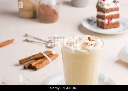Ein Glas Milchpunsch mit Zimtpulver auf geschlagenem Eiweiß gegen ein Kuchenstück auf dem Cafeteria-Tisch auf hellem Hintergrund Stockfoto