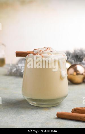 Glas Milchpunsch mit Zimtpulver auf geschlagenen Eiweiß gegen Pinienzapfen am Weihnachtstag auf hellem Hintergrund Stockfoto