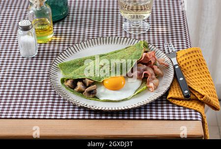 Hausgemachtes Frühstück mit Spinatpfannkuchen mit Speck, Ei und Pilzen auf einem weißen Teller mit einem Salz- und Ölshaker auf einer karierten Tischdecke. Stockfoto