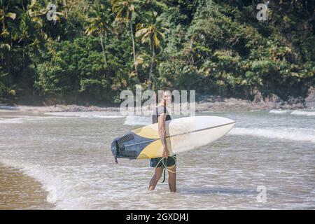 Junger Athlet mit lockigen Haaren und Tattoos in feuchter Sportkleidung, der Surfbrett hält, während er die Kamera auf dem Wasser anschaut Stockfoto