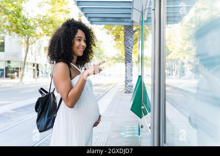 Schwangere afroamerikanische Frau, die den Bauch berührt, während sie Informationen auf dem Display für die Navigation auf der Stadtstraße sucht Stockfoto