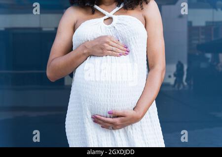 Kurzgeschnittene, nicht erkennbare, schwanger afrikanisch-amerikanische Frau, die ein weißes Kleid in der Nähe einer reflektierenden Wand trägt Stockfoto