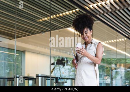 Angenehme, schwanger afroamerikanische Frau mit lockigen Haaren, Textnachrichten auf dem Mobiltelefon, während sie ihren Laptop in der Nähe des Glasgebäudes trug Stockfoto